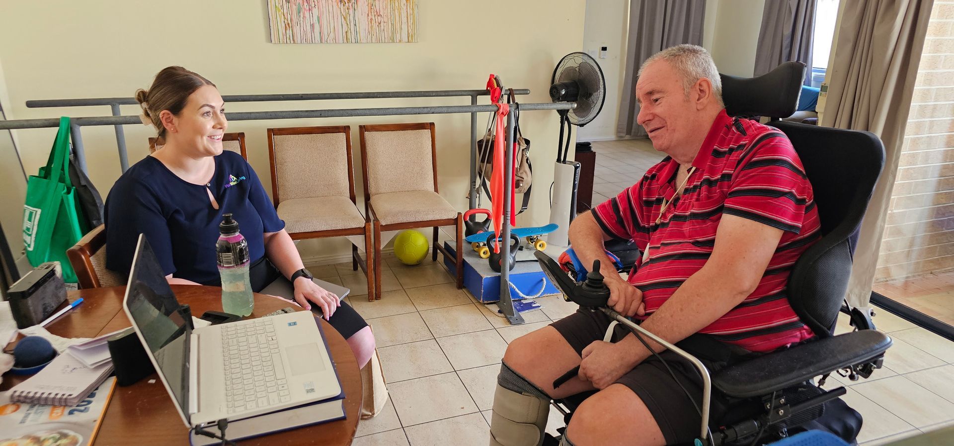 A man in a wheelchair is sitting at a table talking to a nurse.
