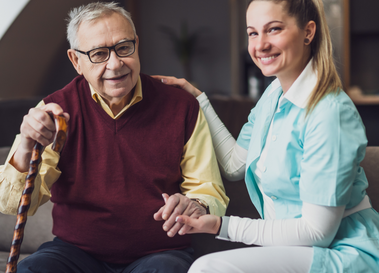 An elderly man with a cane is sitting on a couch with a nurse.