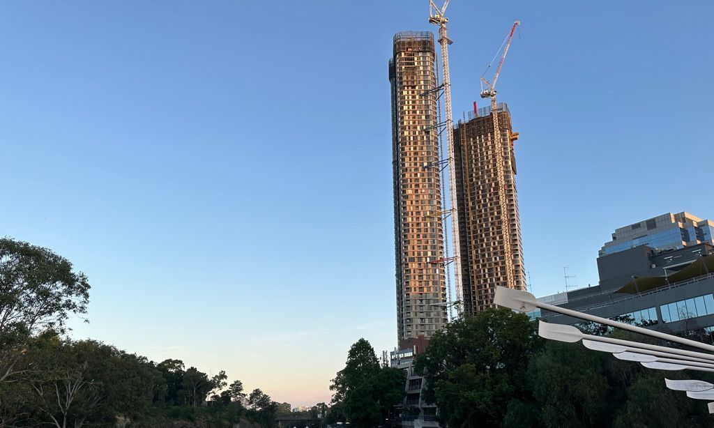 Two tall buildings are being built in a city with trees in the foreground.