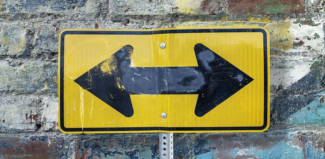 Yellow sign with a black arrow pointing both directions; mounted on a brick wall.