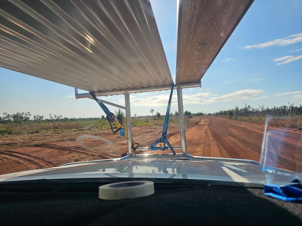 View from inside a vehicle, looking forward. Roof panels secured on roof rack. Dirt road, blue sky, and scrubland — KNG Roofing And Construction in Darwin, NT