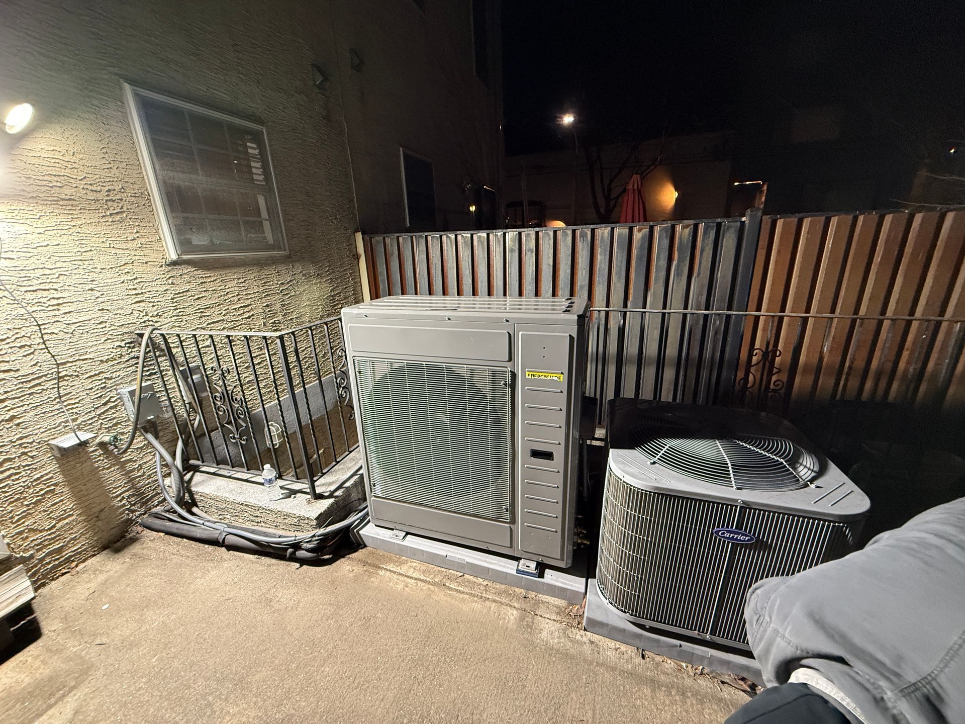 An air conditioning unit and other equipment sit outside at night, next to a building and fence.
