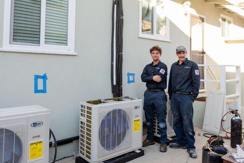 Two HVAC technicians standing next to an air conditioning unit outside a building.