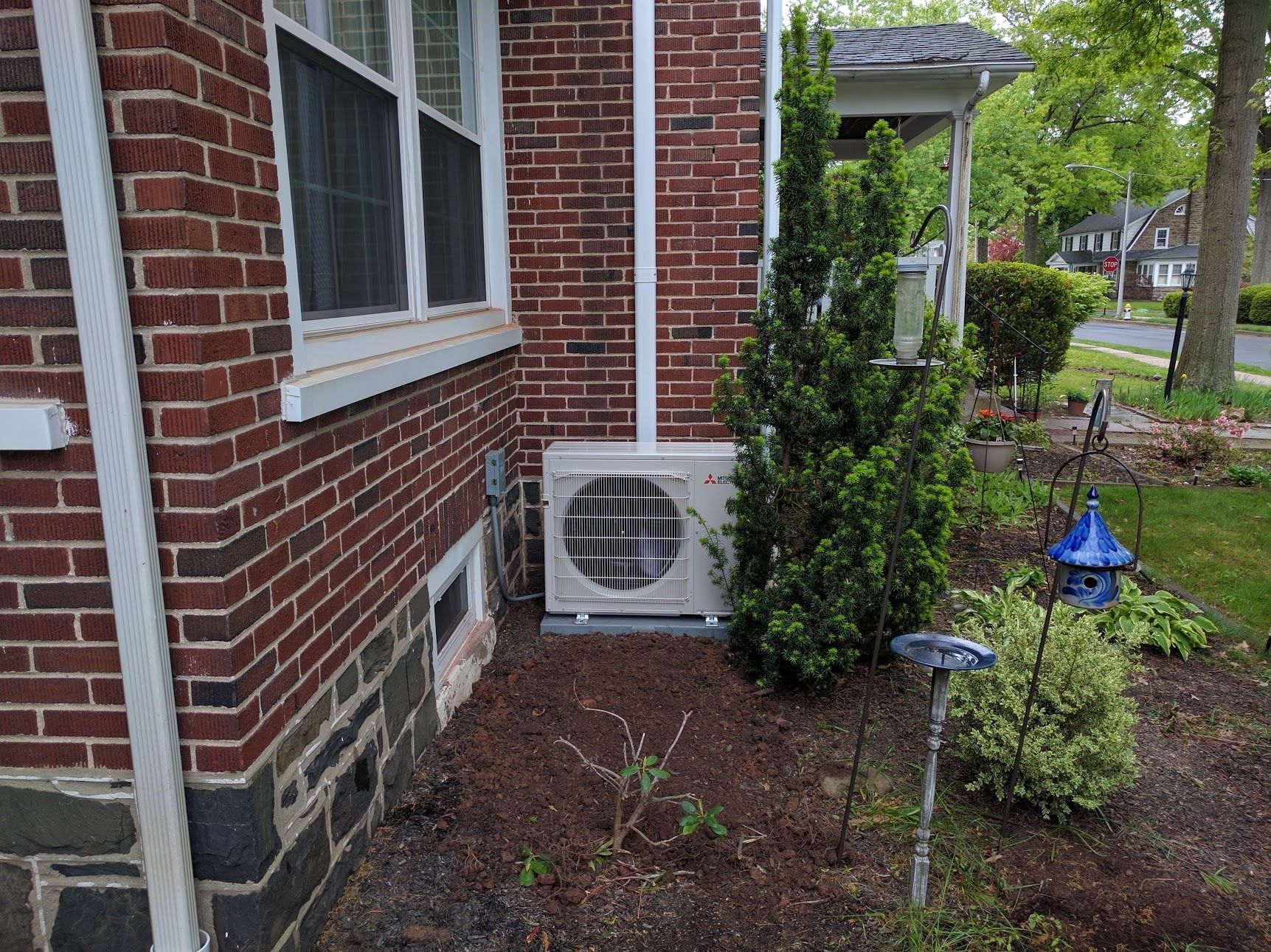 An air conditioning unit sits next to a brick house and a window.