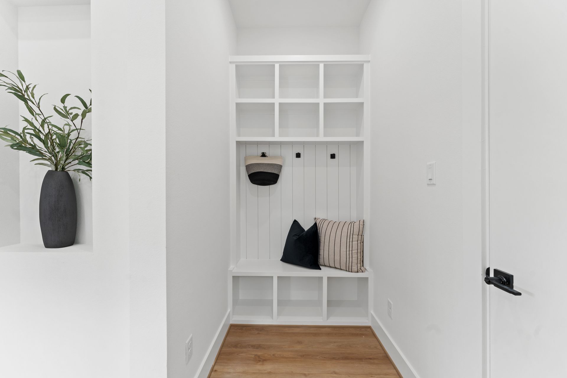 White hallway with built-in storage unit. Wooden floor, black vase with greenery on left.