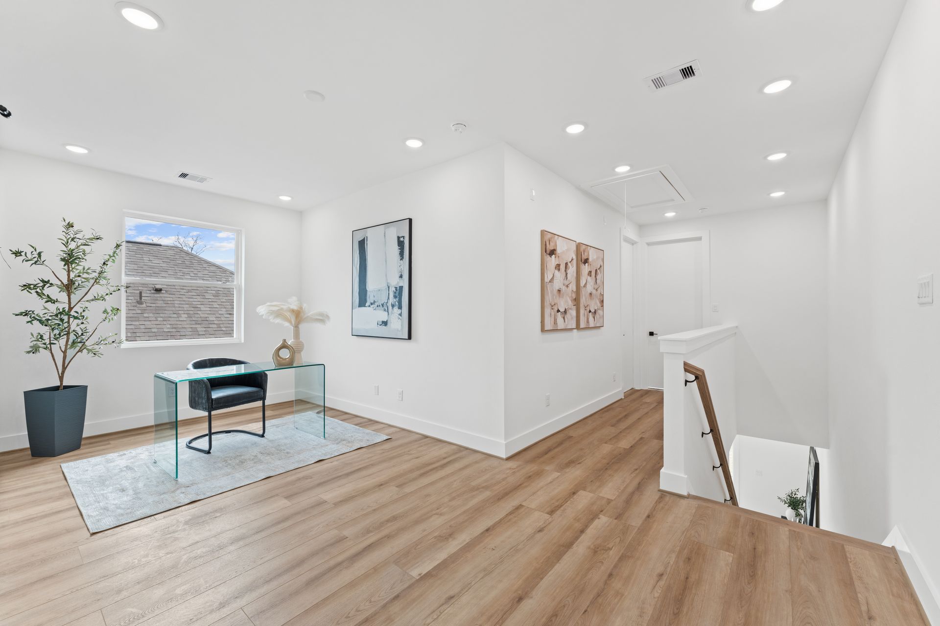 Light-filled home office with a glass desk, art, and a stairway. Pale wood floors and white walls.