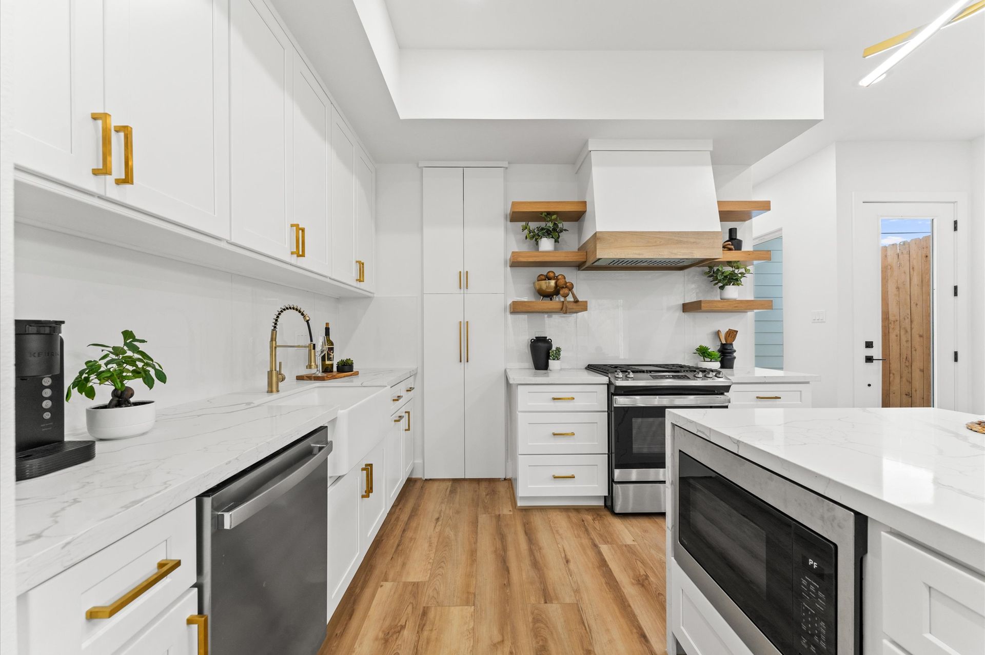 White kitchen with gold hardware, wood shelves, stainless steel appliances, and light wood floors.
