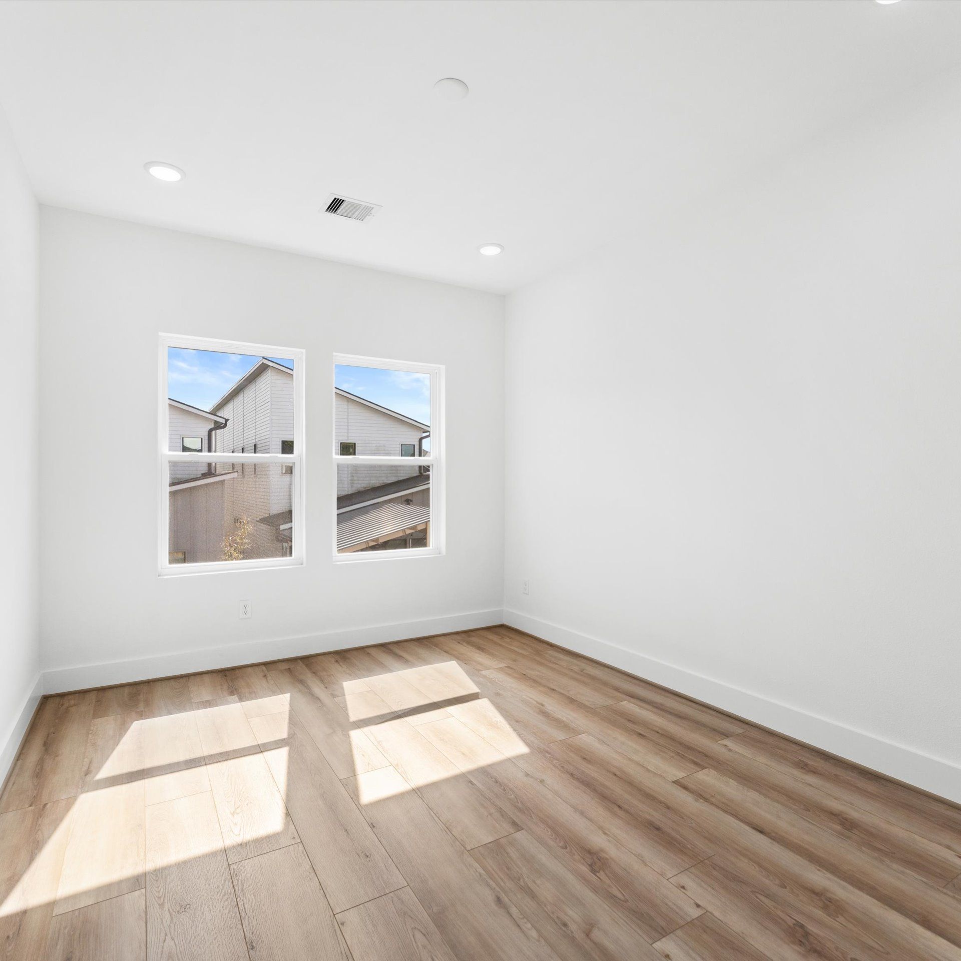 Empty white room with two windows, wood floors, and recessed lighting.