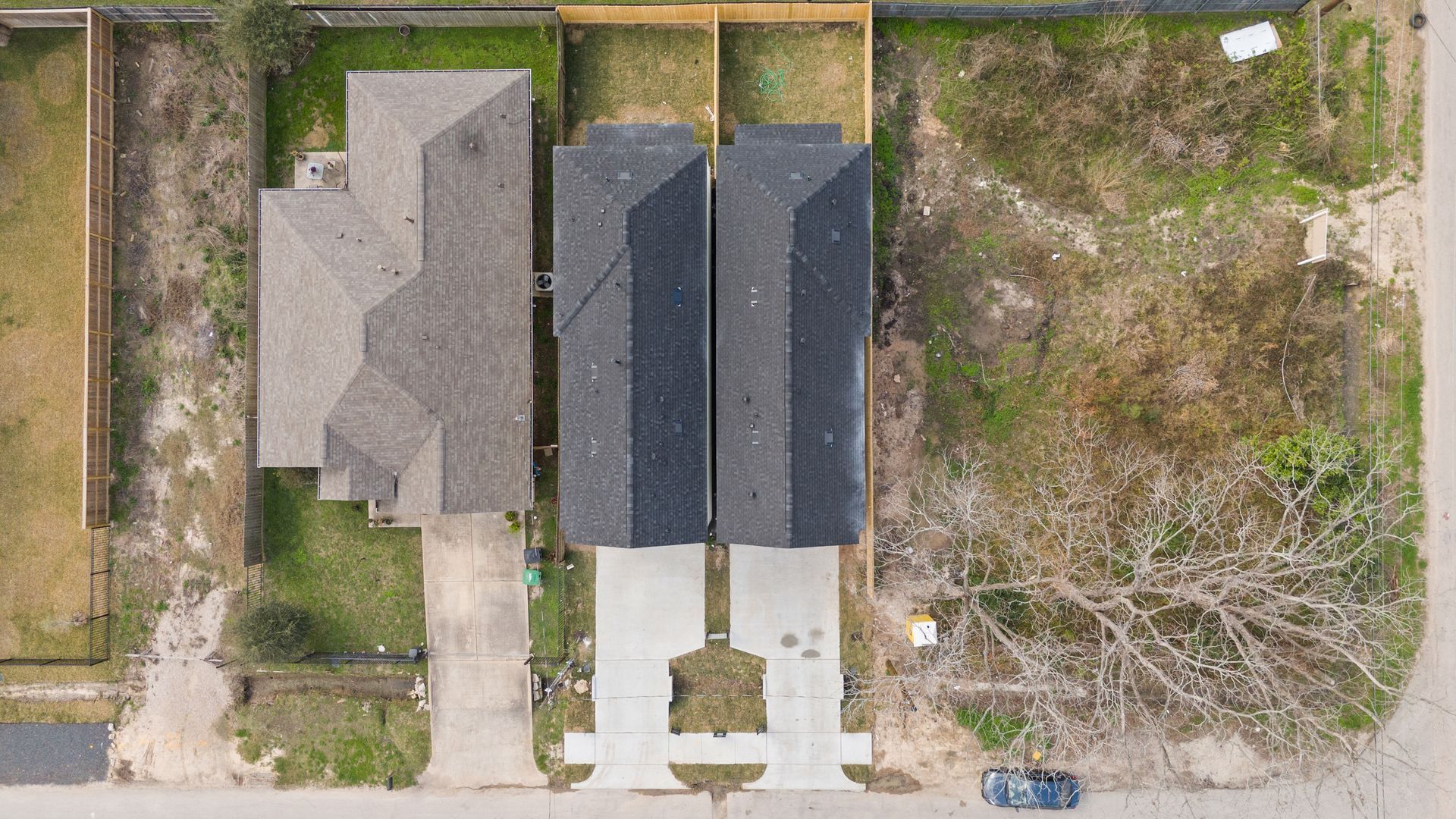 Overhead view of three houses: one tan roof, two black roofs, with driveways and dry grassy yards.