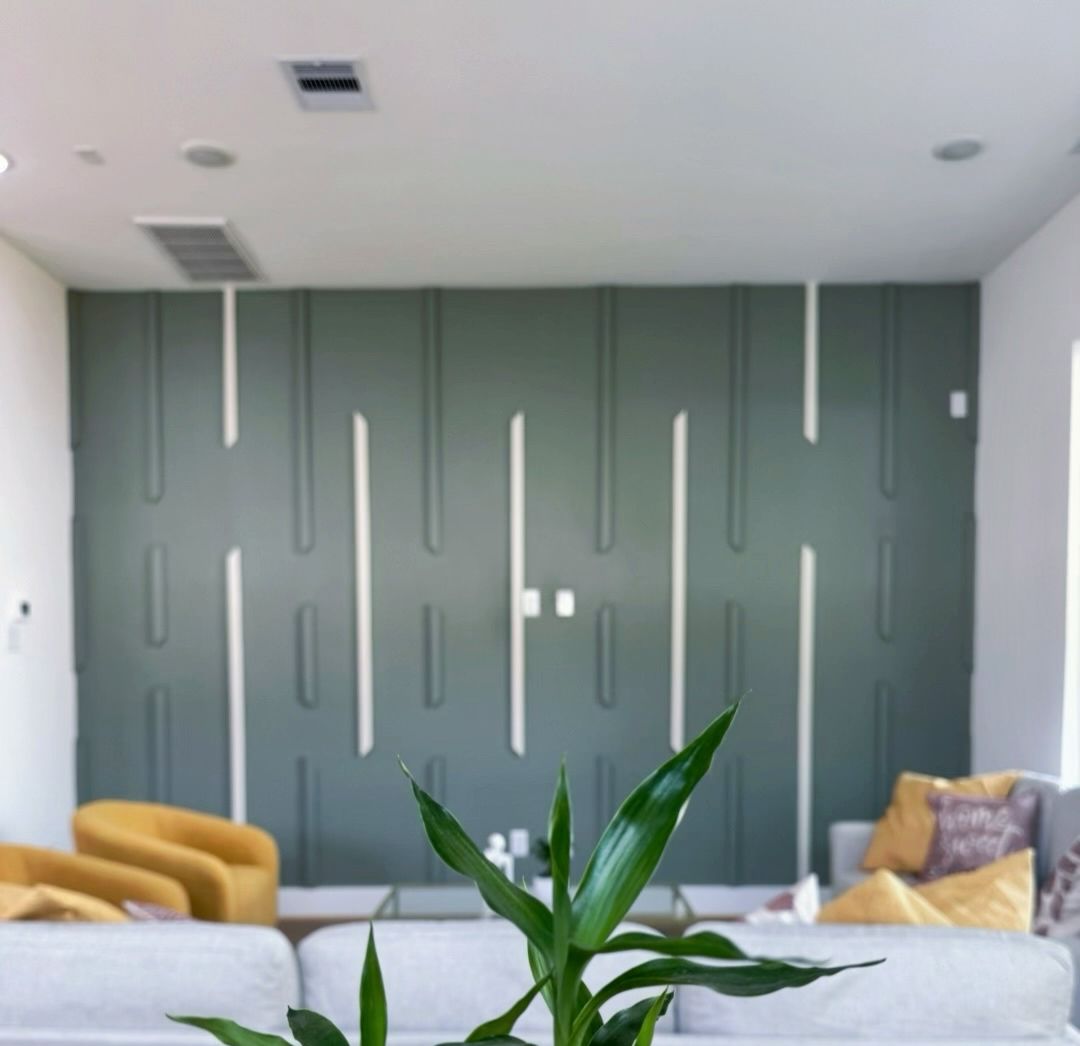 Living room with green accent wall, white trim, yellow and gray sofas, and plant in foreground.
