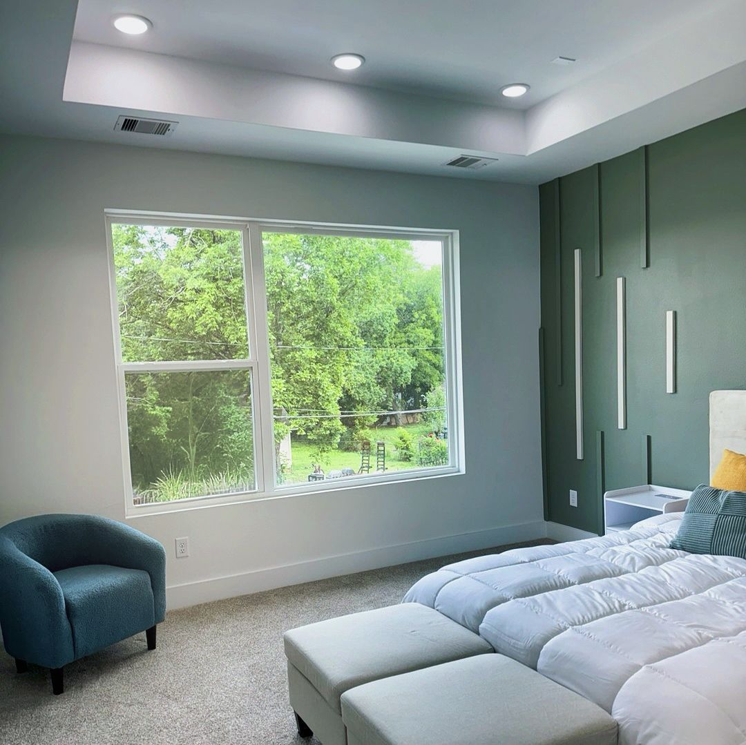 Bedroom with a large window overlooking greenery, blue accent chair, and bed with green accent wall.