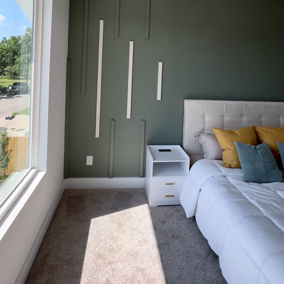 Bedroom with olive-green accent wall, white accents, large window, and a bed with yellow and blue pillows.