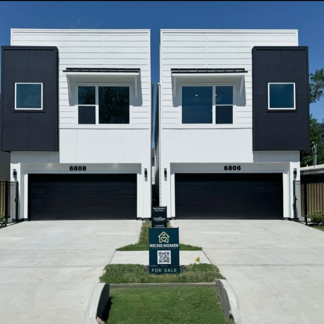 Two modern duplexes, white with black accents, black garage doors, sign in front.