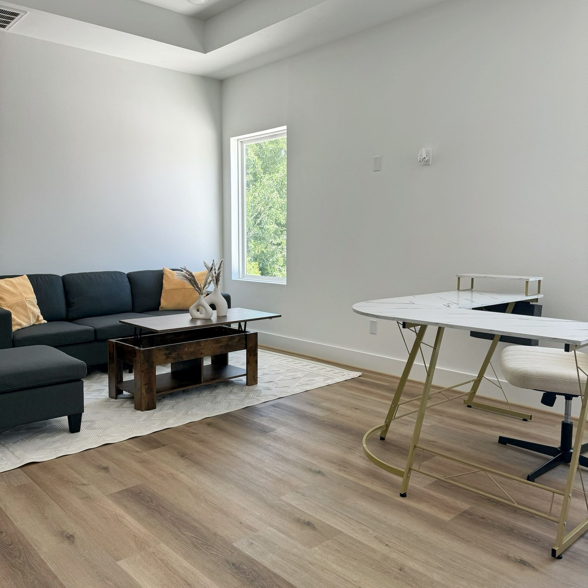 A living room with a sectional sofa, coffee table, desk, and a window with a view.