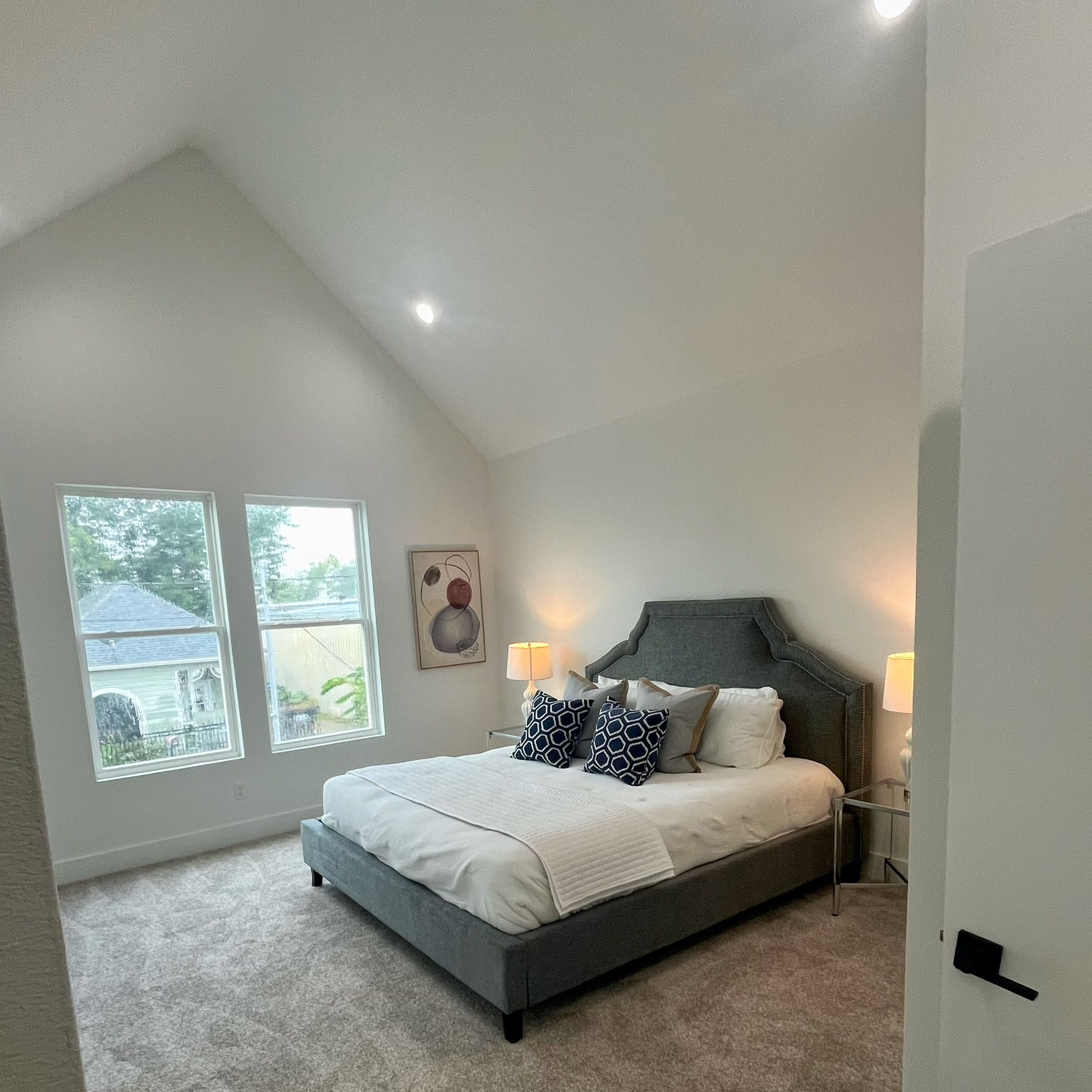 Bedroom with a gray upholstered bed, tall windows, and a vaulted ceiling.