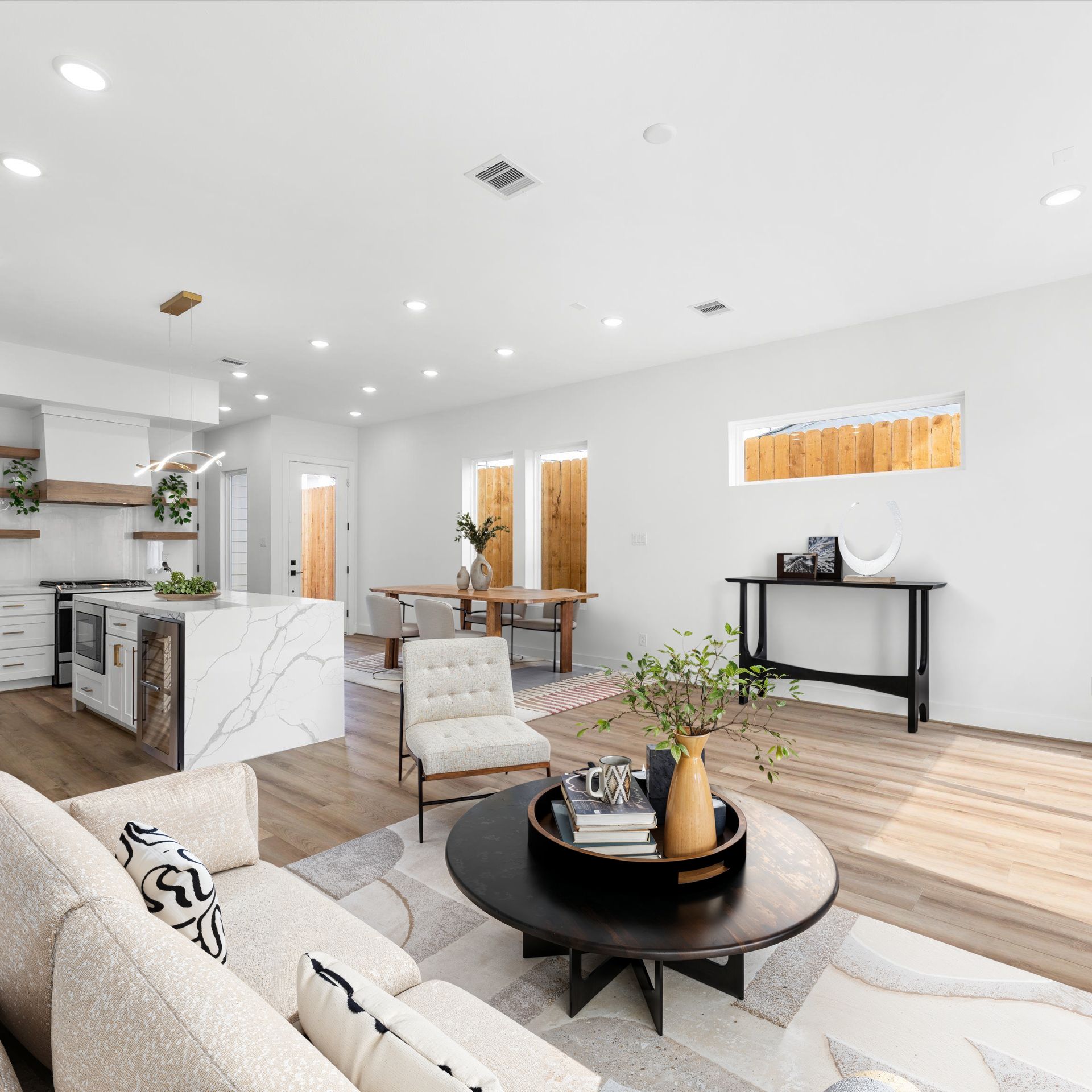 Open-concept living room with kitchen, dining, and seating areas. White walls, light wood floors, and neutral furniture.