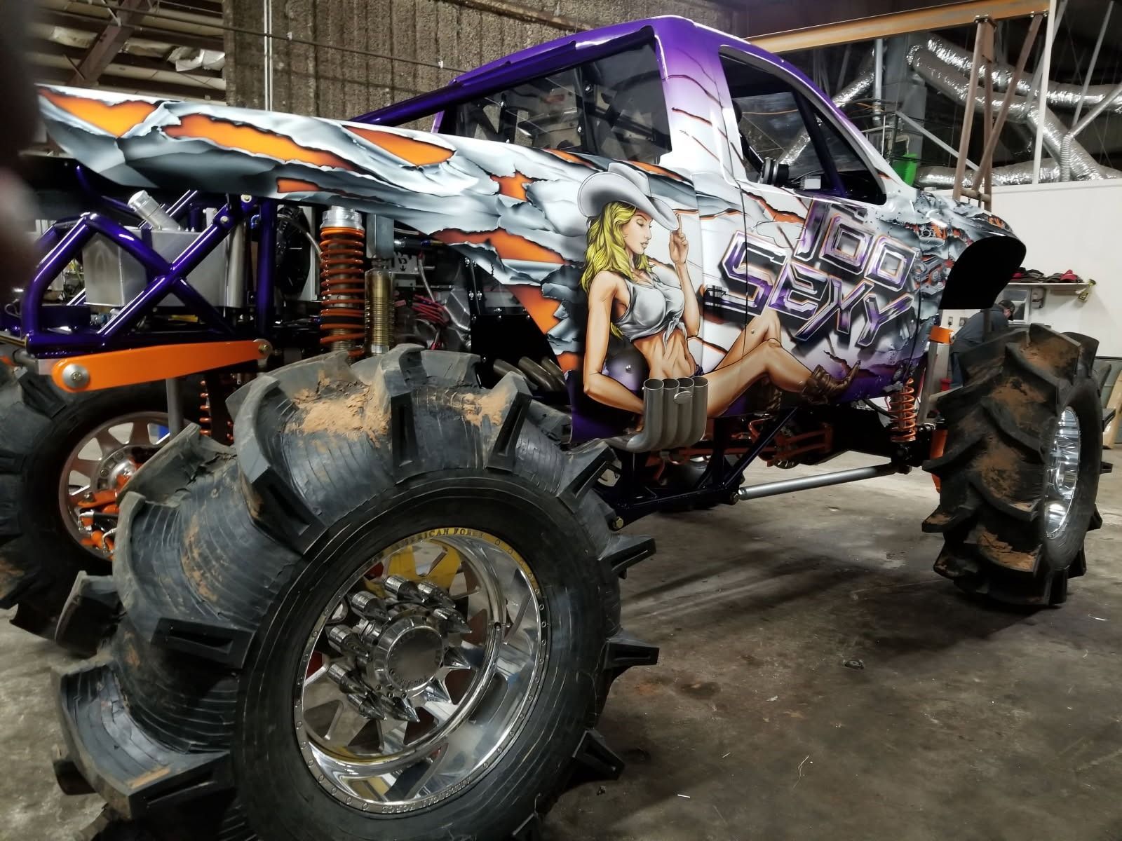 Mud bogging truck with large tires, orange and purple paint, and a pin-up graphic.