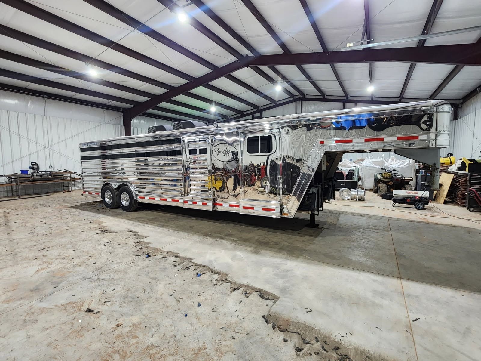 A large, shiny horse trailer inside a workshop. The trailer is silver and black, with a high roof, parked on a concrete floor.
