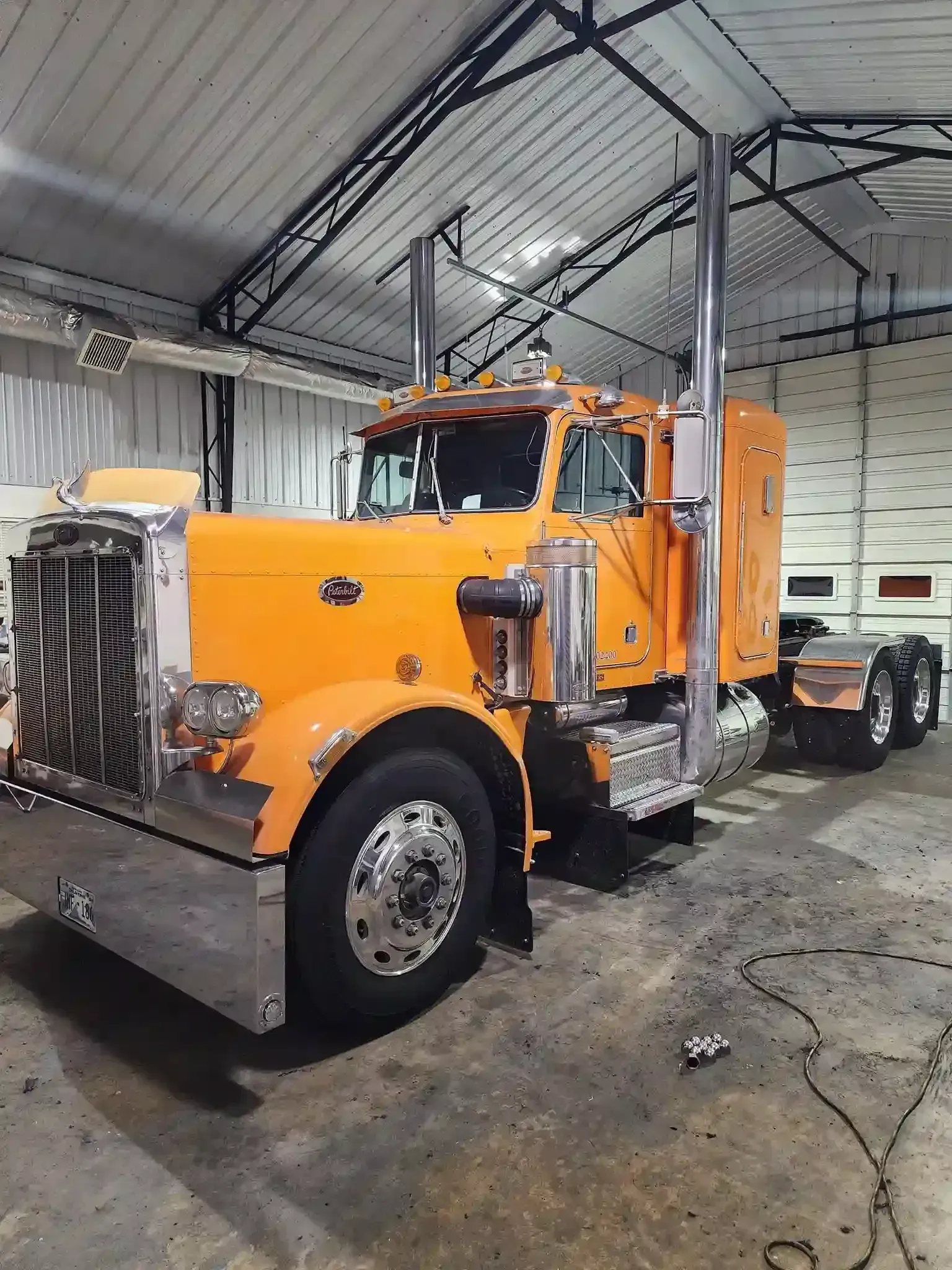 Orange Peterbilt semi-truck parked inside a garage. Chrome accents, tall exhaust stacks, and a shiny bumper.