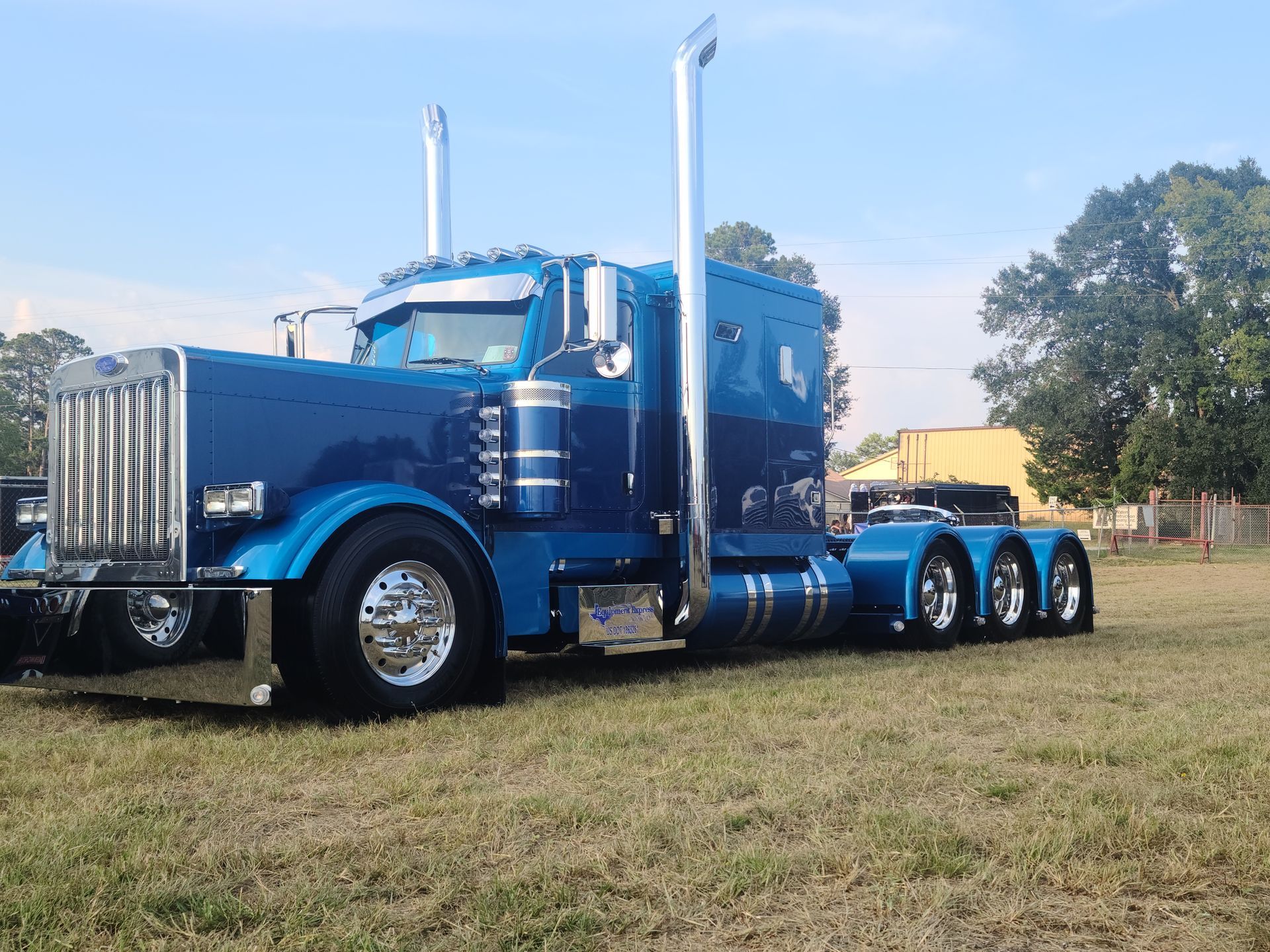 Blue custom Peterbilt semi-truck on grass, chrome accents, bright blue sky.