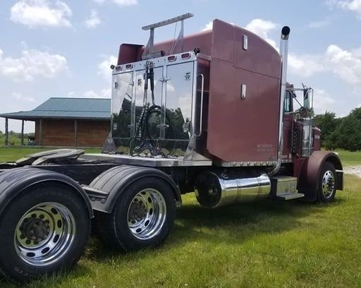 Burgundy semi-truck on grass; polished chrome accents. In background, a wooden building under a blue sky.
