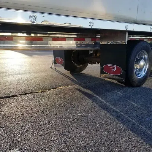 Side view of a semi-trailer with black mud flaps and a shiny chrome rail along the bottom edge, parked on asphalt.