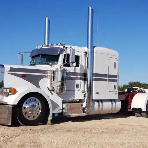White semi-truck with chrome accents and dual exhaust pipes parked on dirt against a blue sky.