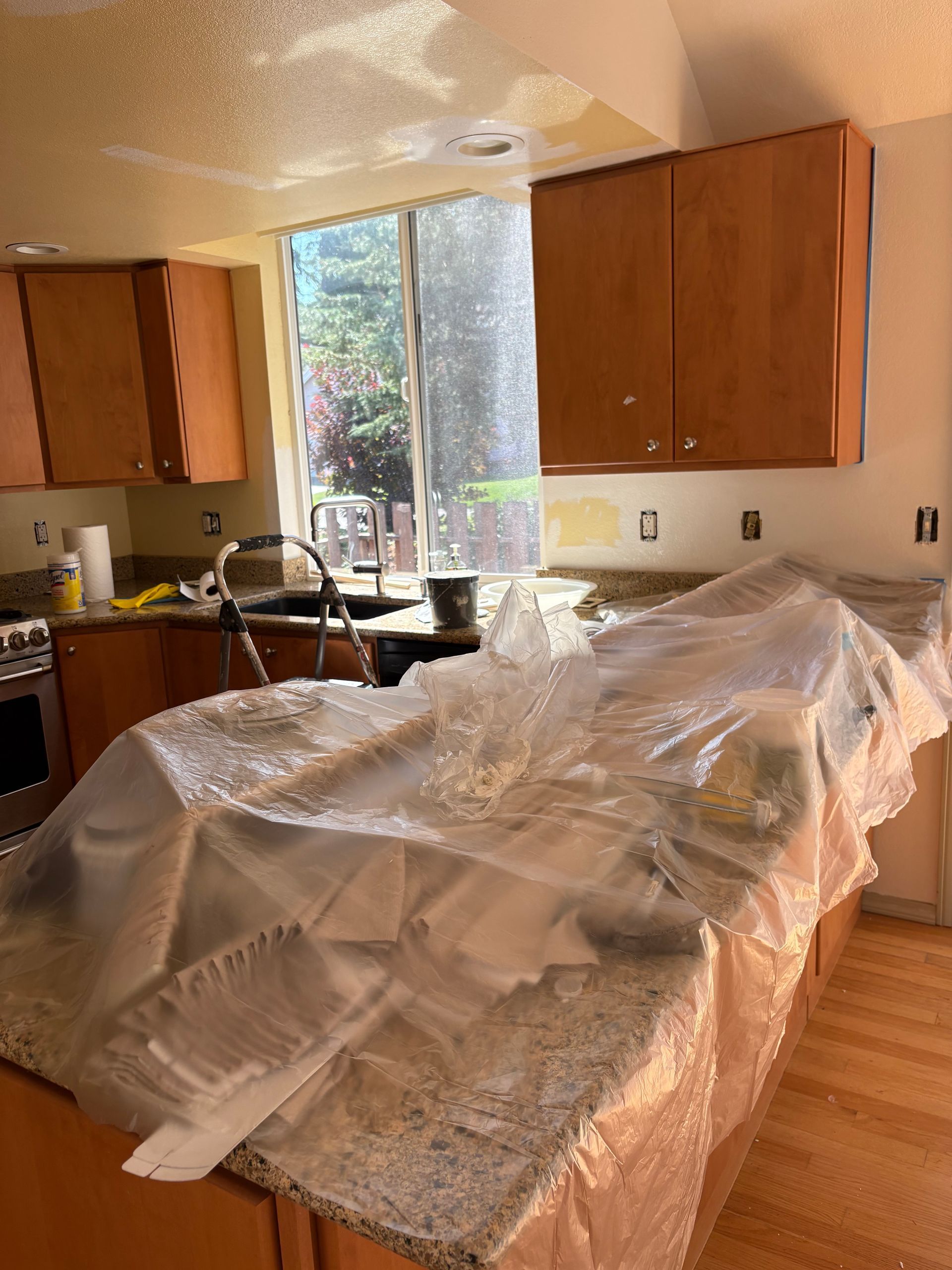 Kitchen painting in progress: countertops covered with plastic, cabinets, window, partially painted walls.