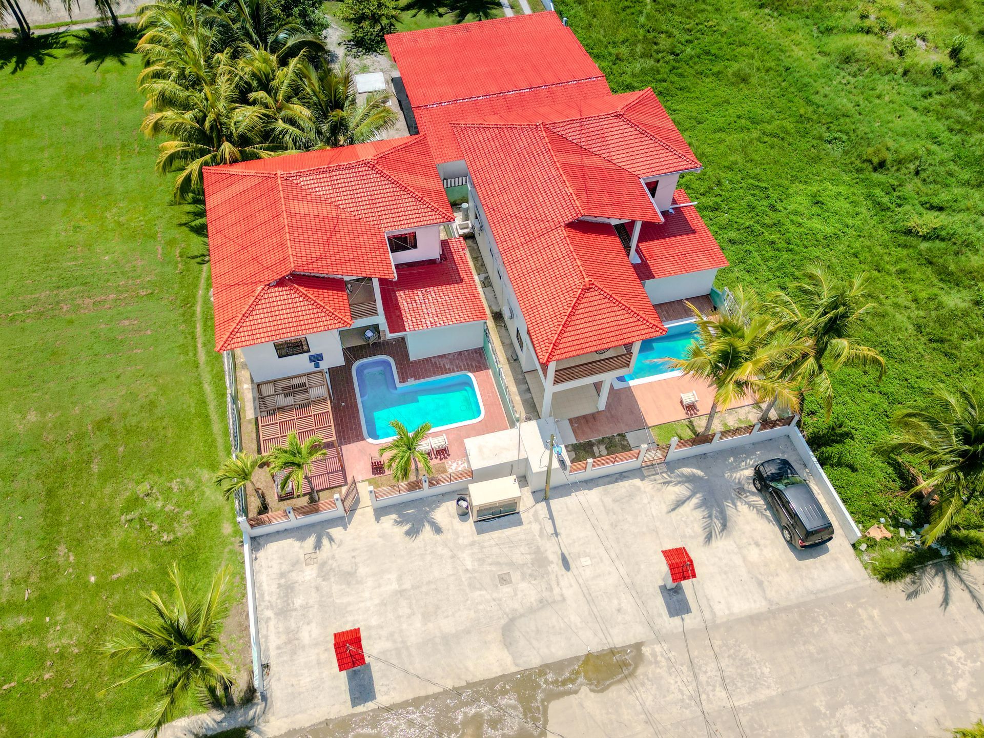 Vista aérea de dos casas blancas con techos rojos y piscinas, sobre un lote de concreto, rodeadas de césped verde.