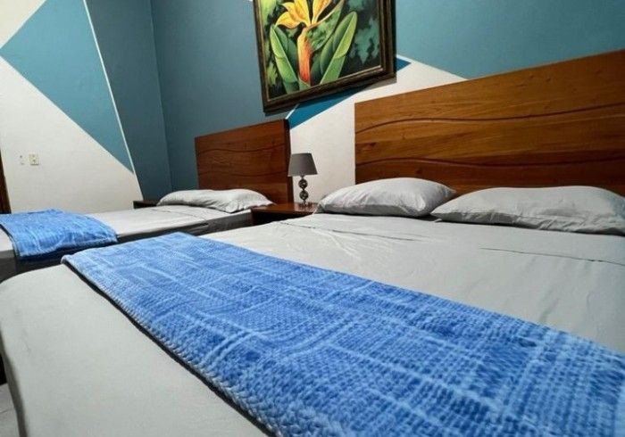 Dormitorio con dos camas con cabecero de madera: una queen y otra individual. Cubrecamas azules y un cuadro.