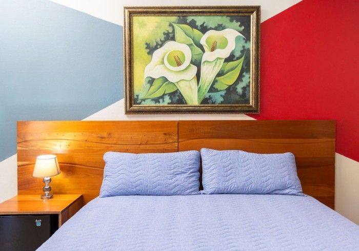 Dormitorio con cabecero de madera, ropa de cama azul y un cuadro de flores blancas. Diseño de pared en rojo, blanco y azul.