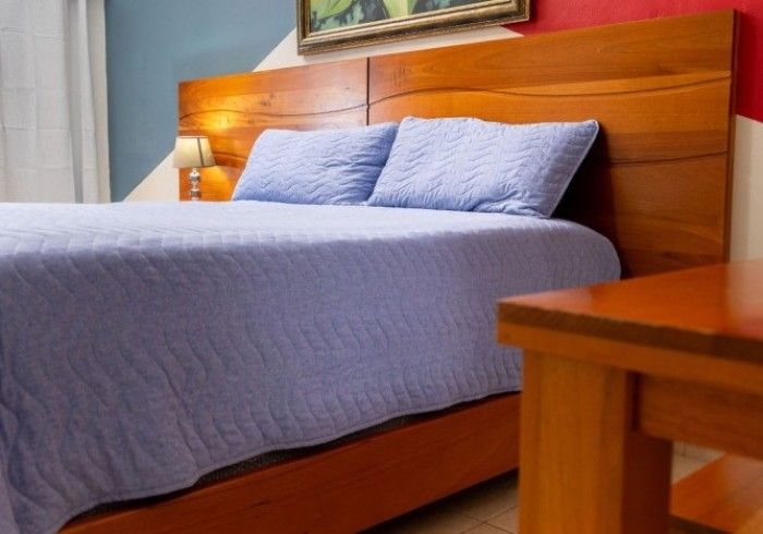 Cama con ropa de cama azul y estructura de madera, mesita de noche a la derecha, paredes rojas y azules.