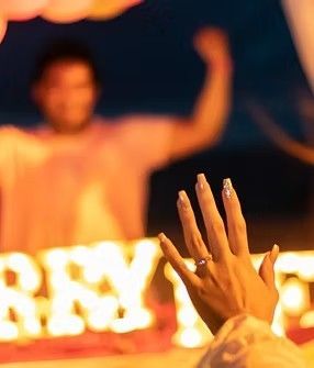 Mano con anillo de compromiso alzada, hombre borroso al fondo. Letrero iluminado que dice