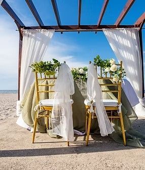 Montaje de boda en la playa: dos sillas doradas con velos blancos, mesa cubierta, vegetación, dosel de madera, cielo azul.