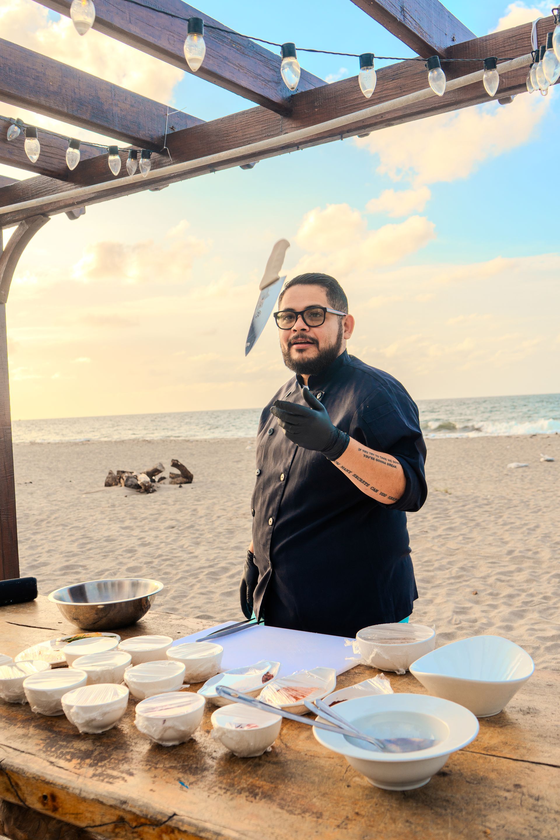 Chef con cuchillo en la playa, preparando comida. Mesa de madera con cuencos, fondo de playa y cielo.
