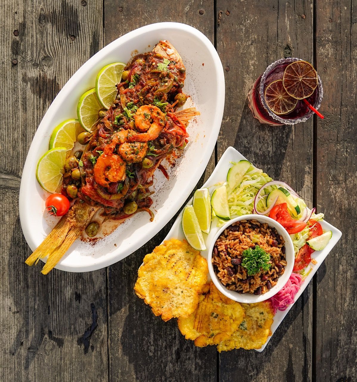 Pescado entero frito con camarones, arroz, tostones, ensalada y cóctel en mesa de madera.