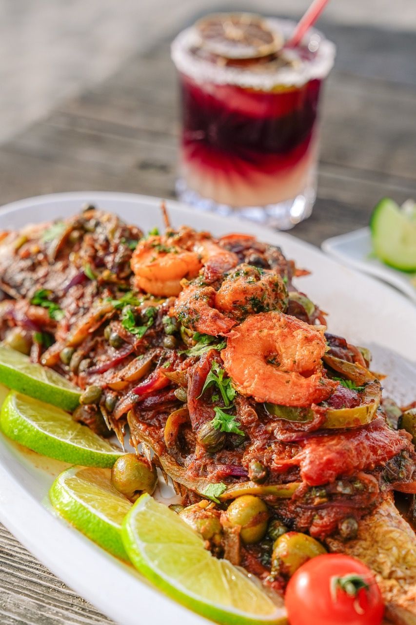 Plato de pescado con camarones, aceitunas y tomates, adornado con limas, servido con un cóctel.