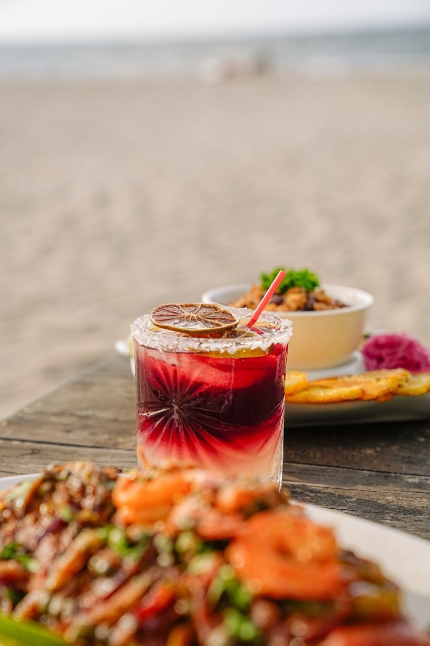 Cóctel con una rodaja de naranja deshidratada, una pajita roja y comida en una mesa de madera en la playa.