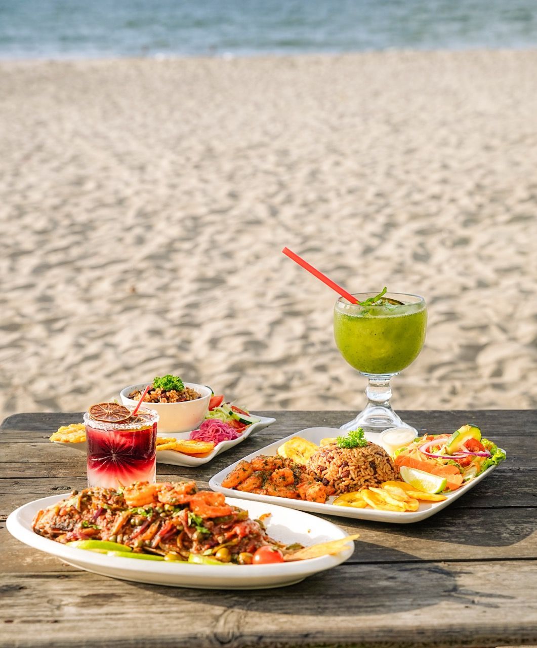 Platos de comida y bebidas en una mesa de madera en la playa: pollo frito, ensalada y bebidas verdes y rojas.