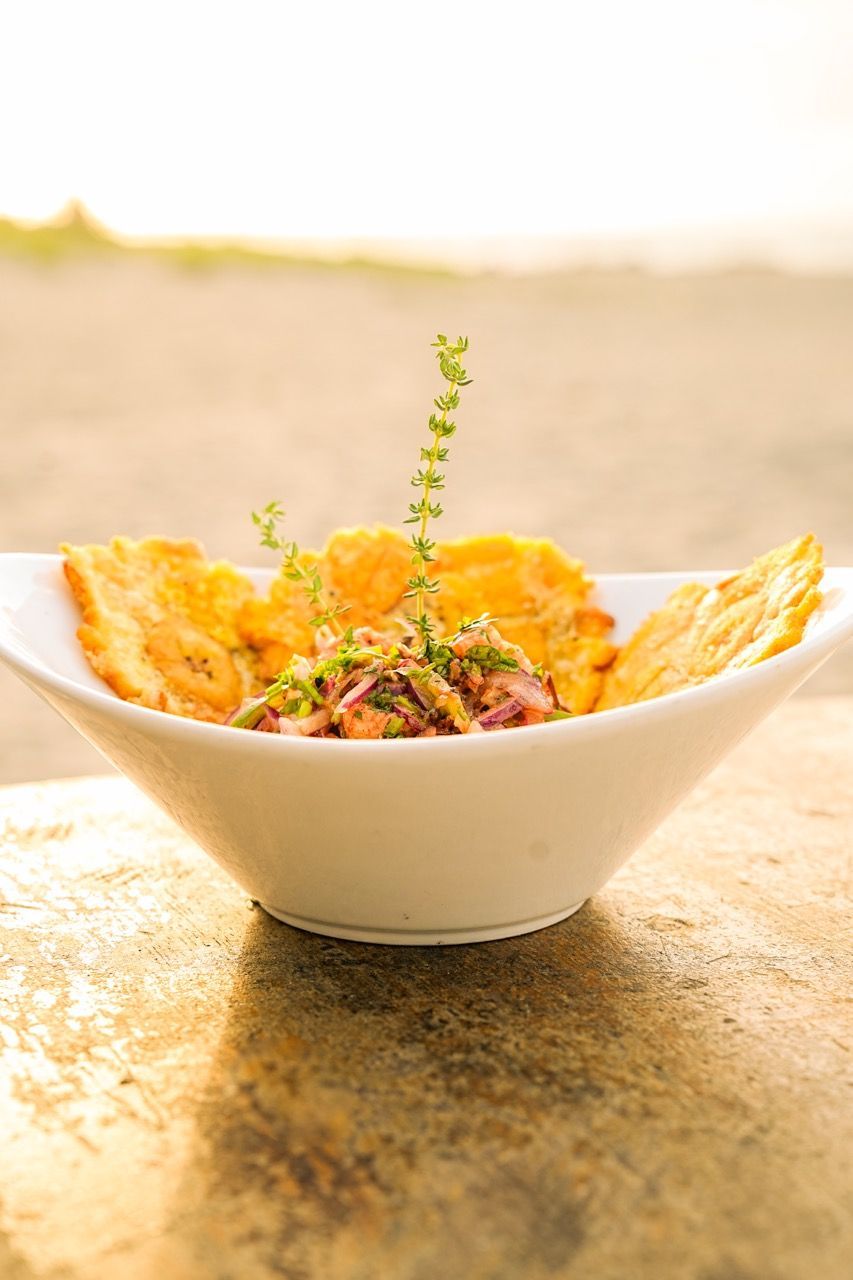 Tazón de comida blanco, chips de plátano que rodean ceviche de mariscos, cubierto con una ramita de hierbas, sobre la mesa.