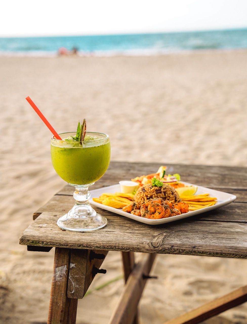 Bebida verde y plato de mariscos sobre una mesa rústica en la playa, con el océano de fondo.