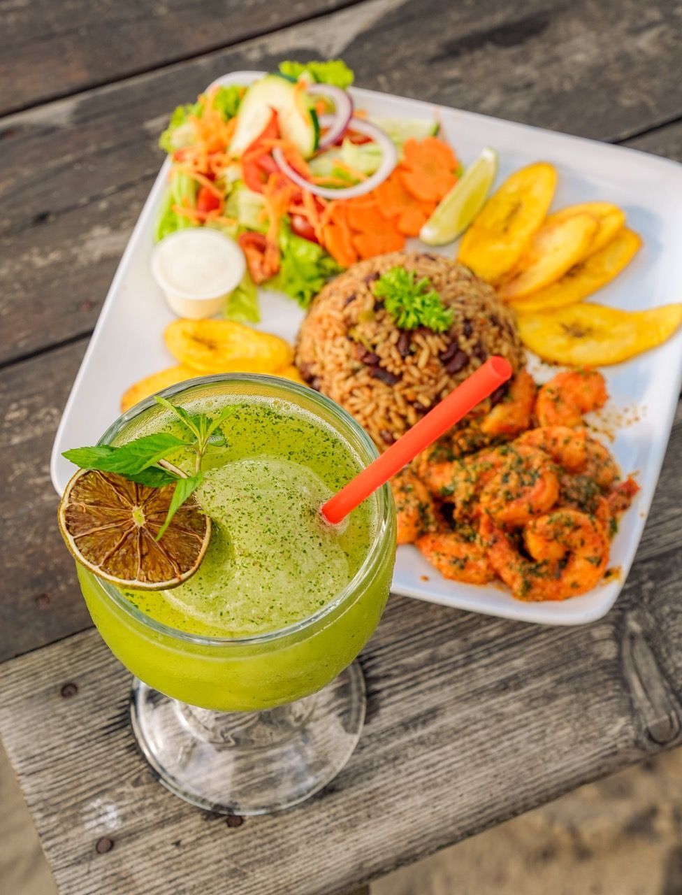 Plato de comida con camarones, arroz, ensalada y plátanos, servido con una bebida verde.