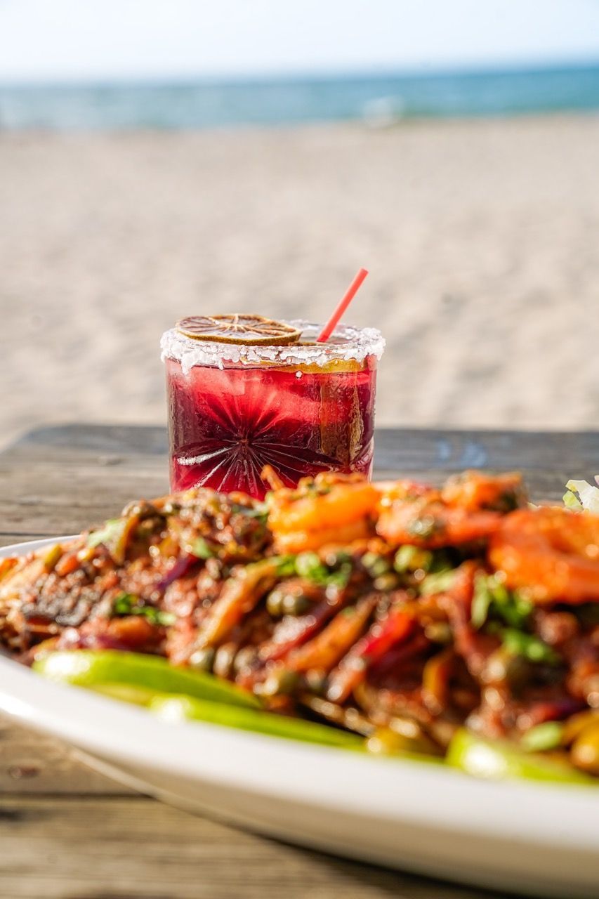 Cóctel y plato de comida sobre una mesa de madera, con fondo de playa. Bebida roja, camarones y ambiente playero.