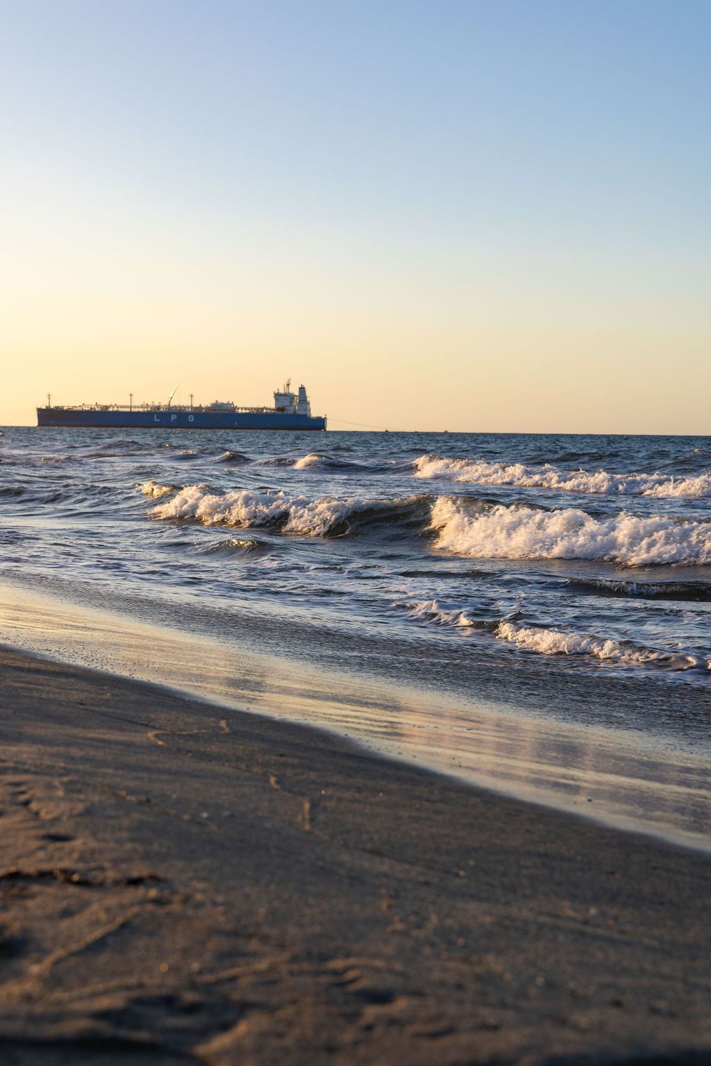 Olas rompiendo en una playa de arena con un barco de carga en el horizonte durante la puesta de sol.