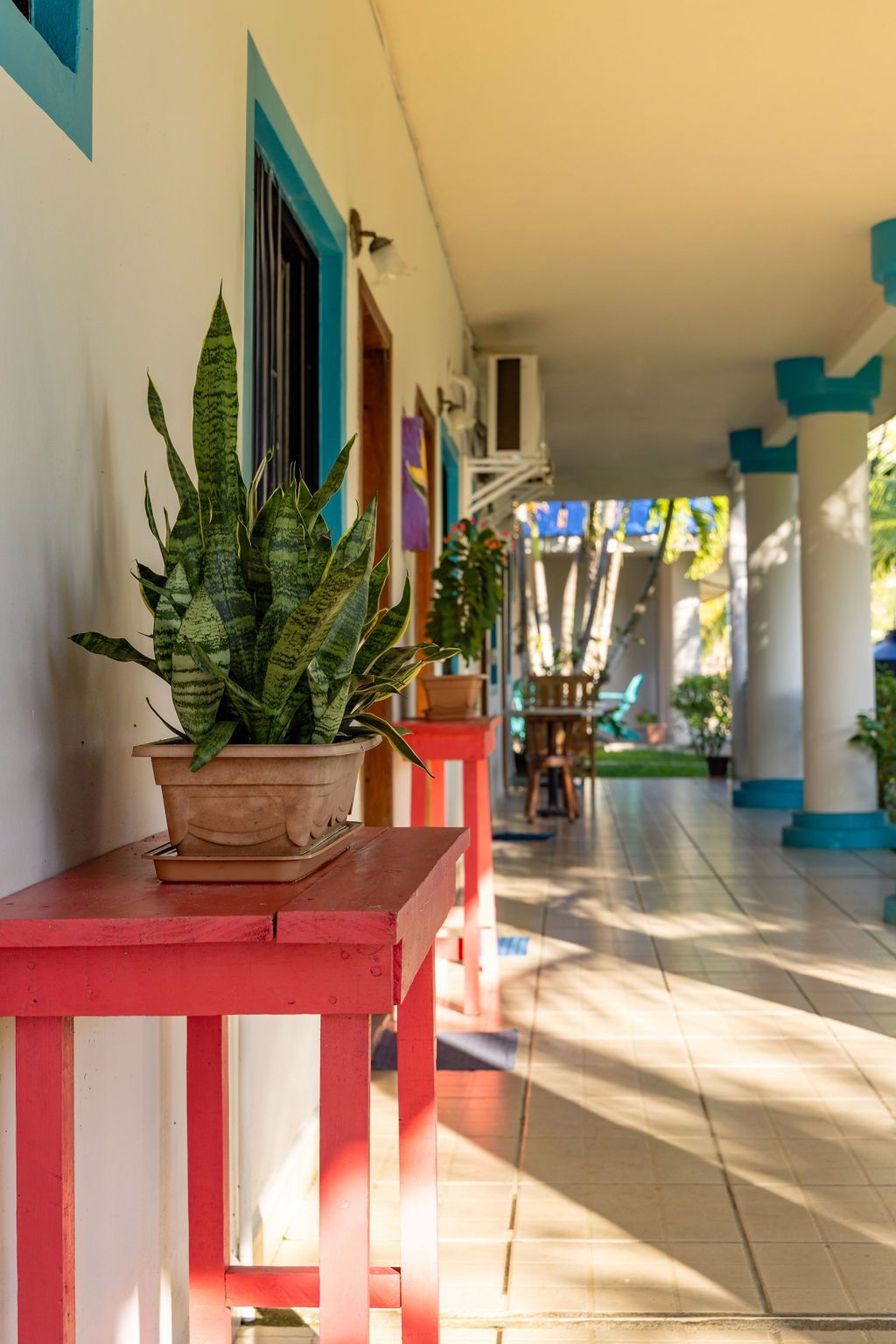 Porche cubierto con mesa roja que sostiene una planta de serpiente en maceta; detalles verde azulado en pilares y puertas.