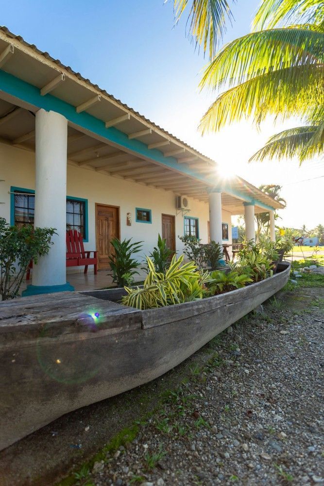 Edificio blanco con detalles en verde azulado, porche con columnas, silla roja, plantas, viejo barco de madera.