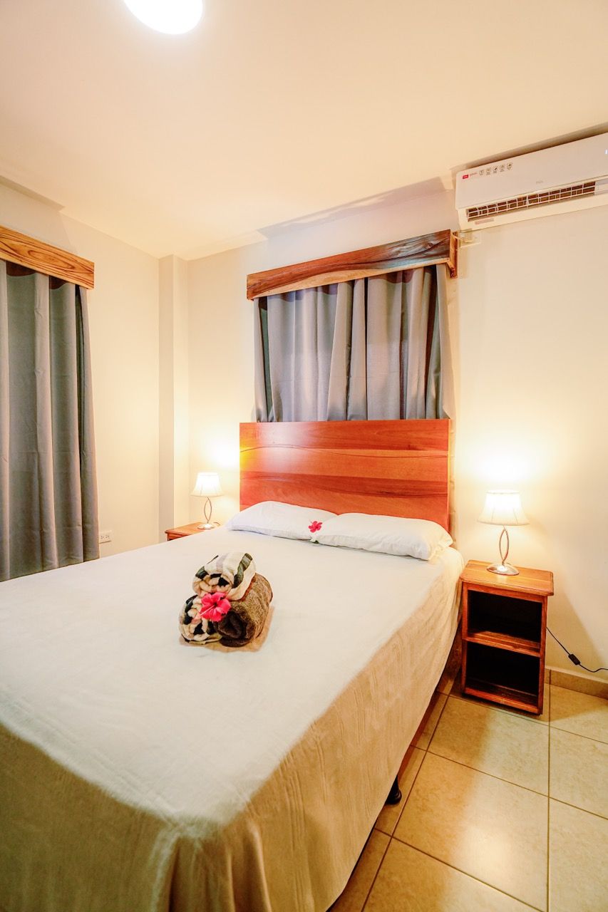 Dormitorio con cama, mesitas de noche, ventana con cortinas y aire acondicionado.