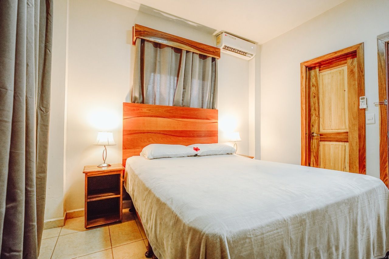 Dormitorio con cama, mesita de noche, cabecero de madera, puerta, cortinas y lámparas.