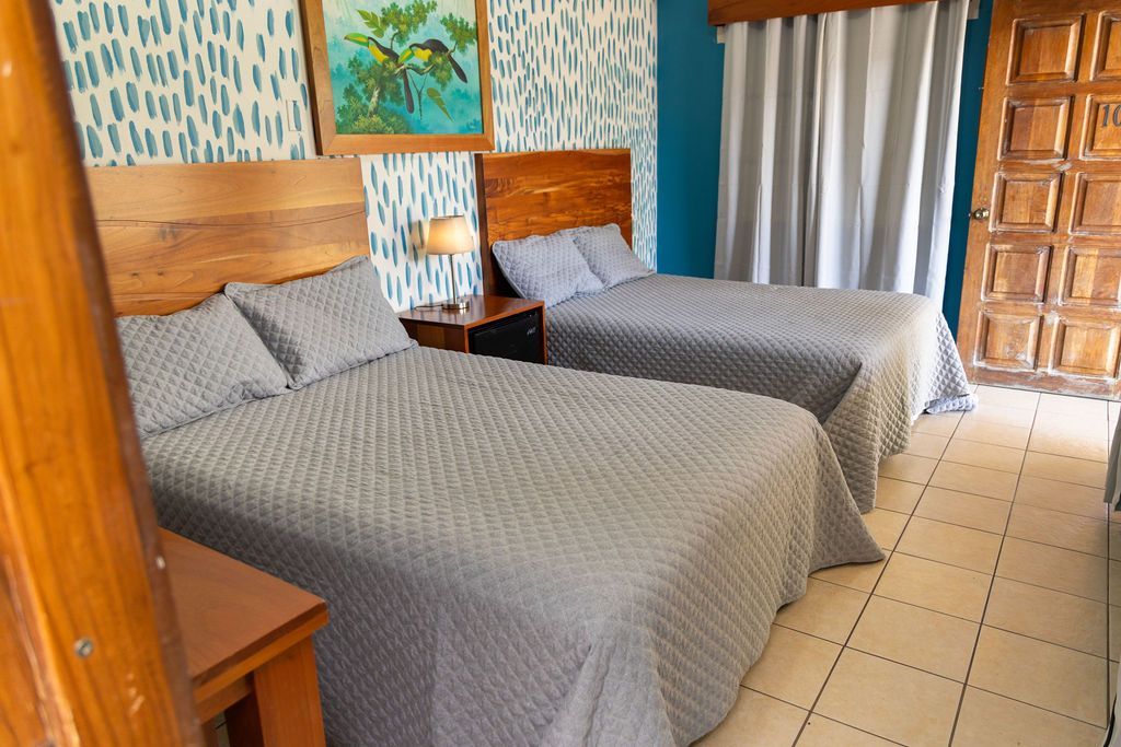 Dormitorio de hotel con dos camas, cabeceros de madera, ropa de cama gris claro, pared verde azulado