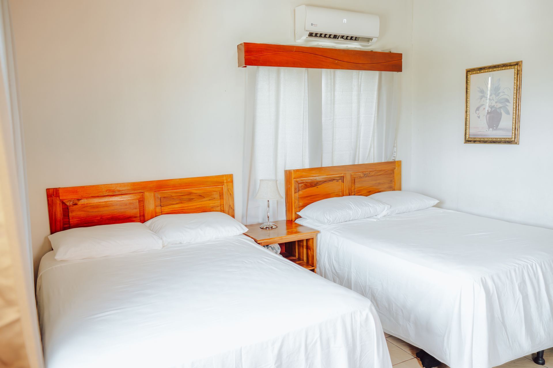 Dos camas en una habitación, ropa de cama blanca, cabeceras de madera, mesita de noche, arte enmarcado y aire acondicionado.